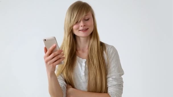 Juventud moderna. hermosa chica de pelo largo de aspecto europeo con el pelo rubio expresivamente hablando en un teléfono inteligente de comunicación de vídeo cerca de la pared blanca — Vídeos de Stock