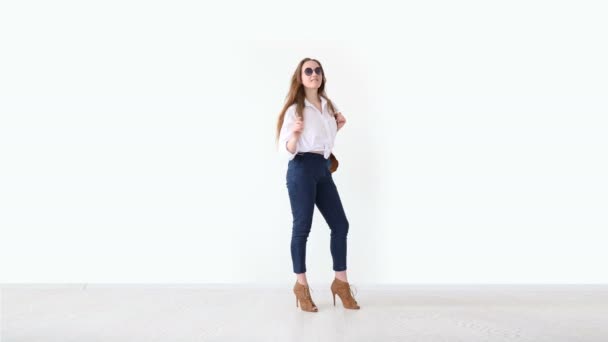 Moda de la juventud moderna. chica elegante posando contra la pared blanca en jeans, camisa blanca, con mochila de cuero y gafas . — Vídeos de Stock