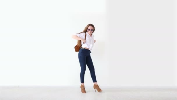 Moda de la juventud moderna. chica elegante posando contra la pared blanca en jeans, camisa blanca, con mochila de cuero y gafas . — Vídeo de stock