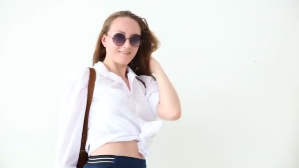 Μόδα της σύγχρονης νεολαίας. κομψό κορίτσι που παρουσιάζουν λευκό τοίχο σε τζην, λευκό πουκάμισο, με δερμάτινο σακίδιο και γυαλιά. — Αρχείο Βίντεο