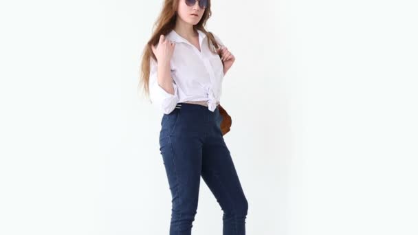 Мода современной молодежи. стильная девушка позирует против белой стены в джинсах, белой рубашке, с кожаным рюкзаком и очками . — стоковое видео
