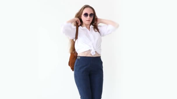 Moda della gioventù moderna. elegante ragazza posa contro muro bianco in jeans, camicia bianca, con zaino in pelle e occhiali . — Video Stock