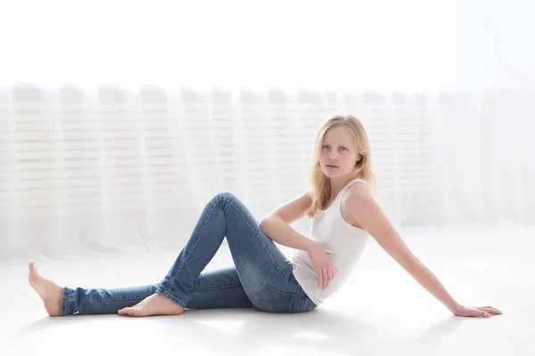 Schöne blonde Frau in Jeans und weißem T-Shirt auf dem Boden in einem hellen Raum — Stockfoto