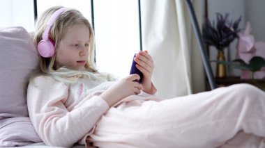 modern life of generation Z. teenage girl in pajamas and headphones in the room on the bed listens to music from a smartphone.