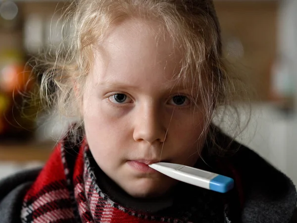 Chica caucásica fría en casa. retrato divertido de un niño enfermo en una bufanda y a cuadros con un termómetro en el sofá en el apartamento — Foto de Stock