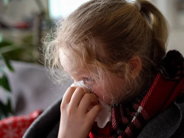 Chica caucásica fría en casa. retrato de un niño enfermo en una bufanda y cuadros en el sofá en el apartamento, la colegiala sopla mocos en una servilleta — Foto de Stock
