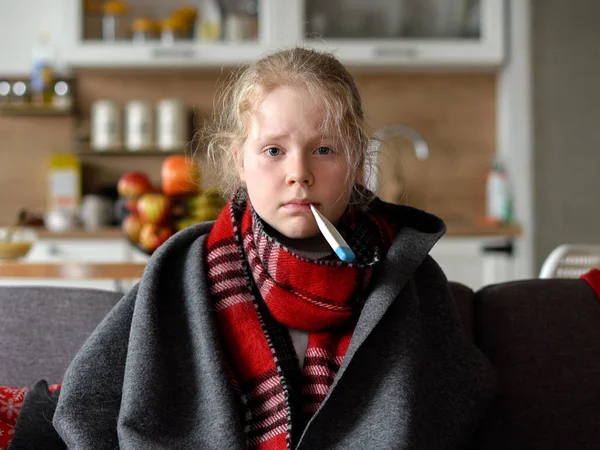 Cold caucasian girl at home. funny portrait of a sick child in a scarf and plaid with a thermometer on the sofa in the apartment — 스톡 사진