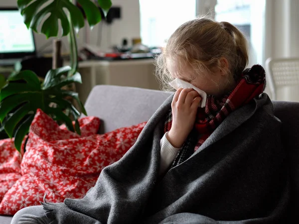 Menina caucasiana frio em casa. retrato de uma criança doente em um lenço e xadrez no sofá no apartamento, a estudante sopra ranho em um guardanapo — Fotografia de Stock