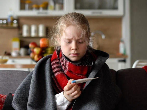 Chica caucásica fría en casa. retrato de un niño enfermo en una bufanda y a cuadros con un termómetro en el sofá en el apartamento —  Fotos de Stock
