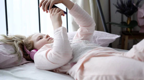 Modern life of generation Z. teenage girl in pajamas and headphones in the room on the bed listens to music from a smartphone. — ストック写真