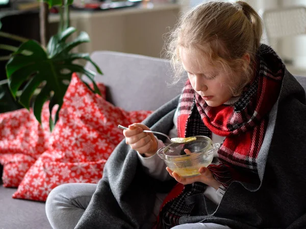 Retrato de un niño enfermo en una bufanda y a cuadros, niña comiendo caldo en el sofá en el apartamento —  Fotos de Stock