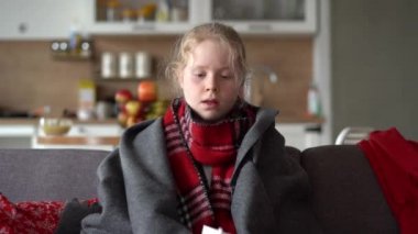 cold caucasian girl at home. portrait of a sick child in a scarf and plaid on the sofa in the apartment, the schoolgirl blows snot into a napkin