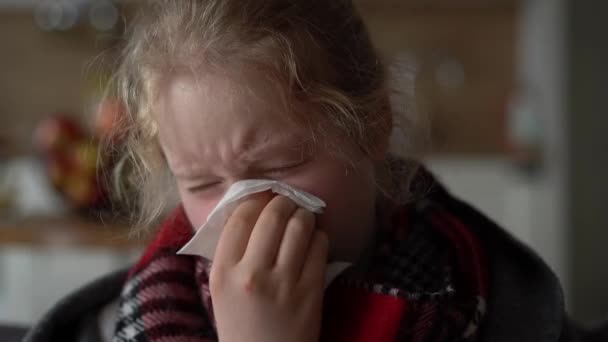 Koud Kaukasisch meisje thuis. portret van een ziek kind in een sjaal en geruit op de bank in het appartement, het schoolmeisje blaast snot in een servet — Stockvideo