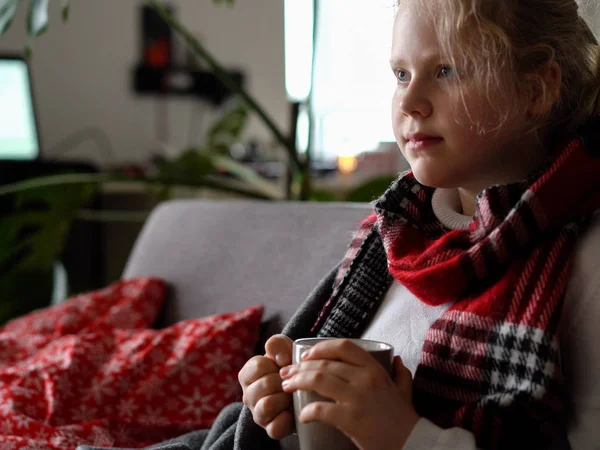 Portrait of a sick child in a scarf and plaid with a cup of hot tea on the sofa in the apartment — Stok fotoğraf