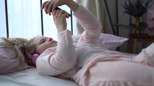 Modern life of generation Z. teenage girl in pajamas and headphones in the room on the bed listens to music from a smartphone. — ストック動画