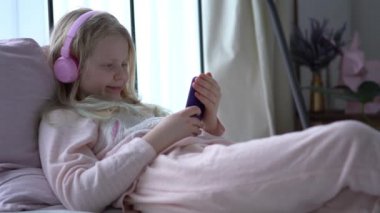 modern life of generation Z. teenage girl in pajamas and headphones in the room on the bed listens to music from a smartphone.