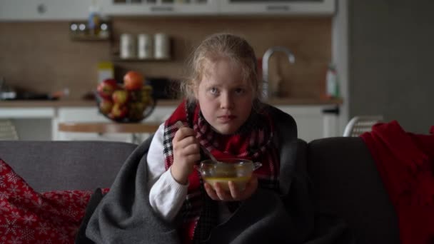 Retrato de un niño enfermo en una bufanda y a cuadros, niña comiendo caldo en el sofá en el apartamento — Vídeo de stock