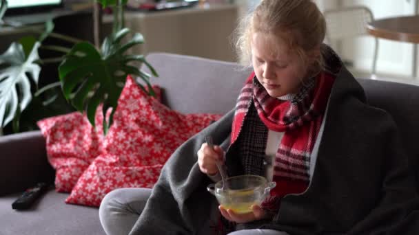 Portret van een ziek kind in een sjaal en plaid, meisje dat bouillon eet op de bank in het appartement — Stockvideo