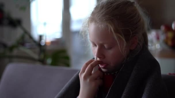 Koud Kaukasisch meisje thuis. portret van een ziek kind in een sjaal en geruit op de bank in het appartement, het schoolmeisje blaast snot in een servet — Stockvideo