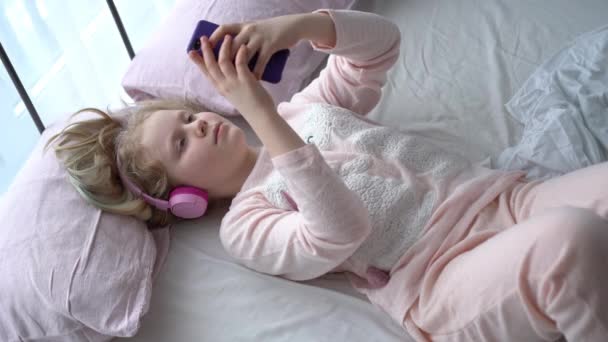Modern life of generation Z. teenage girl in pajamas and headphones in the room on the bed listens to music from a smartphone. — Stok video
