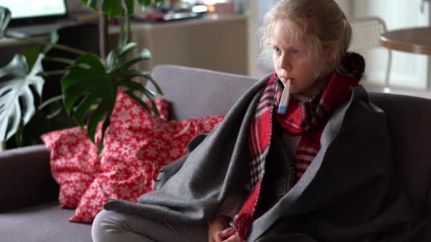 Cold caucasian girl at home. funny portrait of a sick child in a scarf and plaid with a thermometer on the sofa in the apartment — 图库视频影像