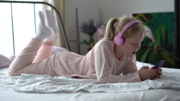 Σύγχρονη ζωή της γενιάς Z. έφηβος κορίτσι με πιτζάμες και ακουστικά στο δωμάτιο στο κρεβάτι ακούει μουσική από ένα smartphone. — Αρχείο Βίντεο