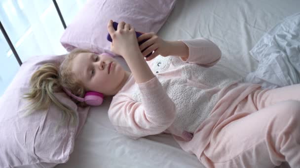 Современная жизнь поколения Z. девочка-подросток в пижаме и наушниках в комнате на кровати слушает музыку со смартфона . — стоковое видео
