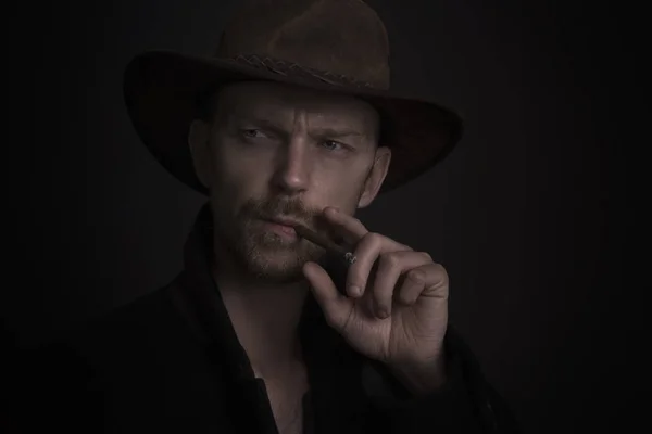 Man wearing hat and smoking a cigar, looking very serious