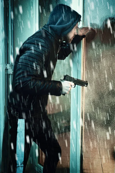 Mysterious hooded man with pistol looking through a window