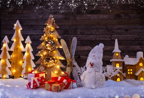 Sfondo natalizio con villaggio in legno illuminato e pupazzo di neve — Foto Stock