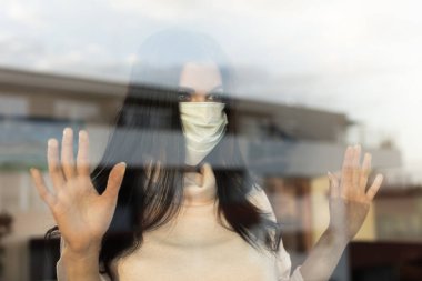 Peçeli genç bir kadın pencereye bakıyor, ev izolasyonu kavramı. Covid-19 salgını evde tek başına. Yeni korona virüsü normal hayatları değiştiriyor.