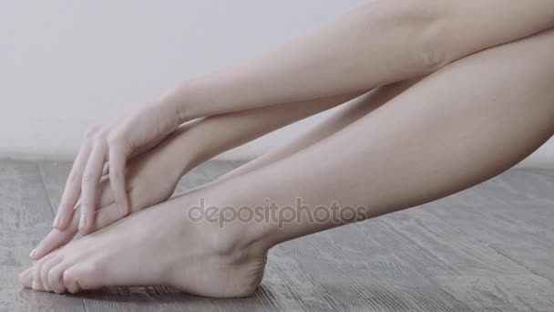 Menina gentilmente tocando suas pernas sentadas no chão de madeira com parede branca no fundo . — Vídeo de Stock