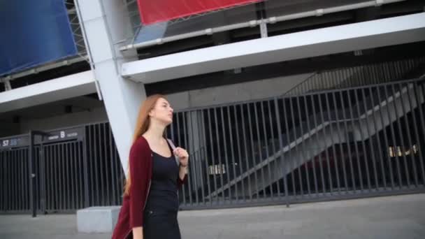 Приваблива дівчина в червоному пальто з червоними губами йде вулицею в місті, ніж перетворюється на камеру і посміхається . — стокове відео