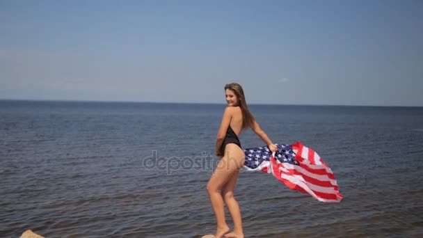 Piękna, szczupła dziewczyna w bikini, cieszyć się życiem pełnym taniec na plaży z flagą Usa — Wideo stockowe