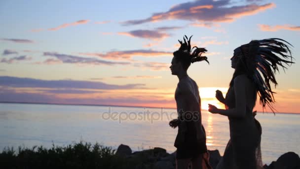 Siluetas de hombre y mujer corriendo corriendo corriendo en la playa al atardecer. Traje y accesorios nativos americanos — Vídeo de stock