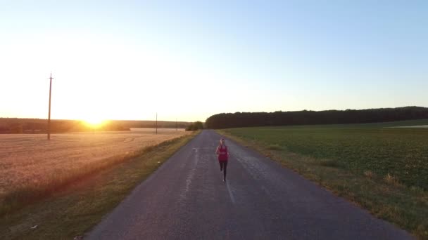 Mujer deportista corriendo una carretera cerca del campo al atardecer . — Vídeo de stock