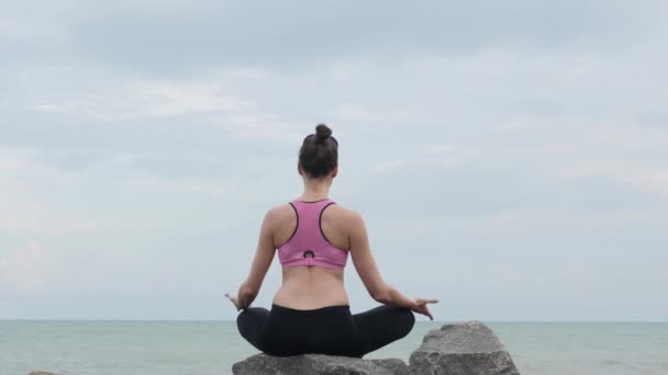 Girl practices yoga fitness exercise near the ocean beach sitting on the stone in lotus pose — Stock Video