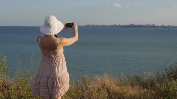 Jonge aantrekkelijke vrouw in mooie jurk kijken naar zonsondergang of zonsopgang Over Bay van de Top van de berg, meisje neemt Panorama foto's op Phone.Hair in de Wind. Handheld atmosferische Shot — Stockvideo