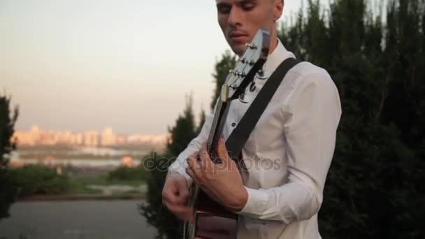Хипстер играет на гитаре и поет на открытом воздухе на живописном закате и зеленом фоне — стоковое видео