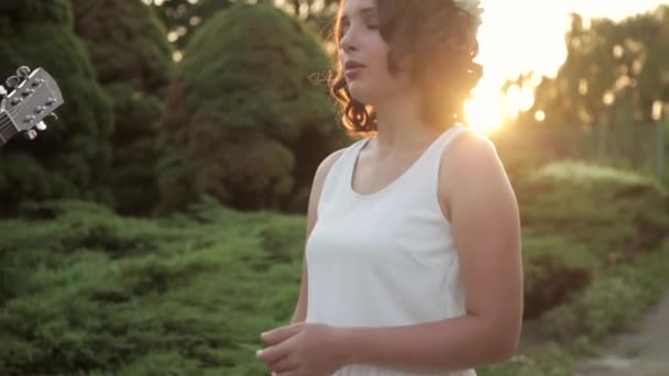 Joven hermosa chica con círculo de flores y vestido blanco canta y baila al aire libre al atardecer — Vídeo de stock