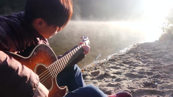 Guapo joven asiático tocando la guitarra en la costa del río bosque al amanecer. Niebla nebulosa al fondo — Vídeo de stock