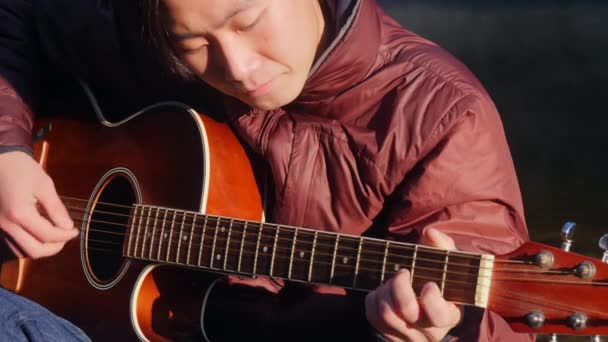 Schöner junger asiatischer Mann, der bei Sonnenaufgang am Ufer des Waldflusses Gitarre spielt. Morgendämmerungsnebel im Hintergrund — Stockvideo