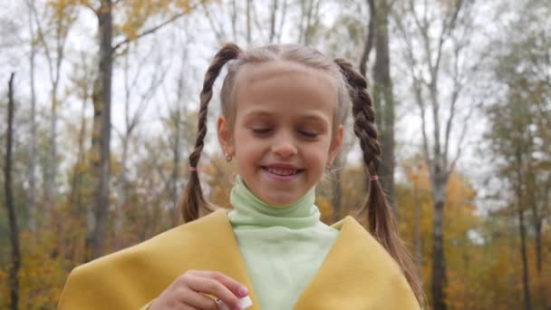 Dziewczynka w żółtym fartuchu dmuchanie baniek powietrza w parku w pochmurny dzień — Wideo stockowe