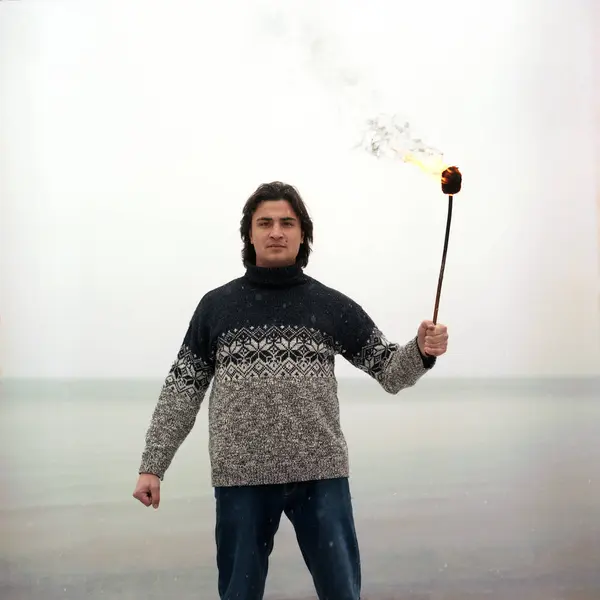 Hombre hispano latino de raza mixta con antorcha de fuego en suéter cálido sobre fondo de mar congelado. Nieve cayendo en primer plano . — Foto de Stock