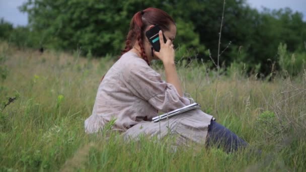 Unga vackra kvinnan sitter på gräset i sommar stadsparken och talar i mobiltelefon. — Stockvideo