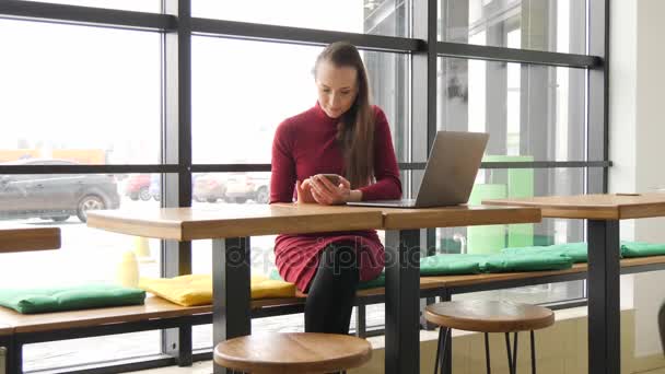 Mujer en vestido rojo usando aplicación en el teléfono inteligente en la cafetería bebiendo café y sonriendo — Vídeo de stock