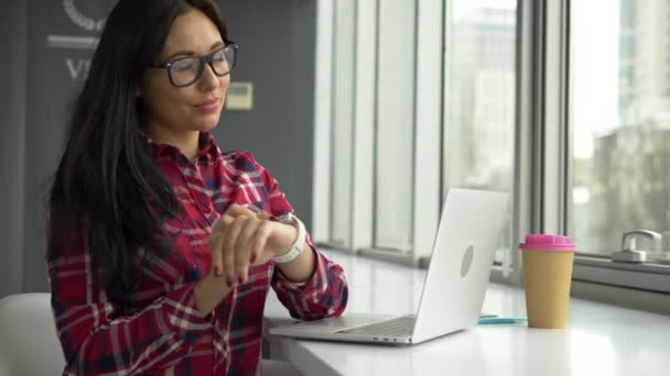 Hipster mujer de raza mixta utilizando smartwatch y trabajando en el ordenador portátil PC en la cafetería — Vídeo de stock