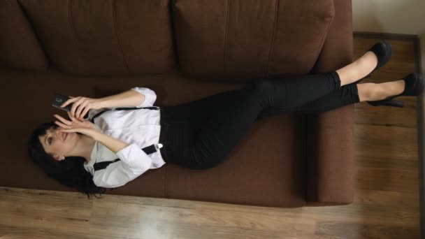 Menina bonita encontra-se em um sofá com telefone celular conversando sms navegar na internet — Vídeo de Stock