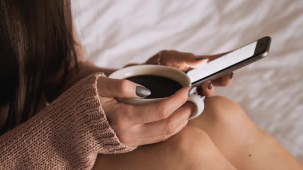 Женщина в постели пьет кофе и проверяет смартфон — стоковое видео