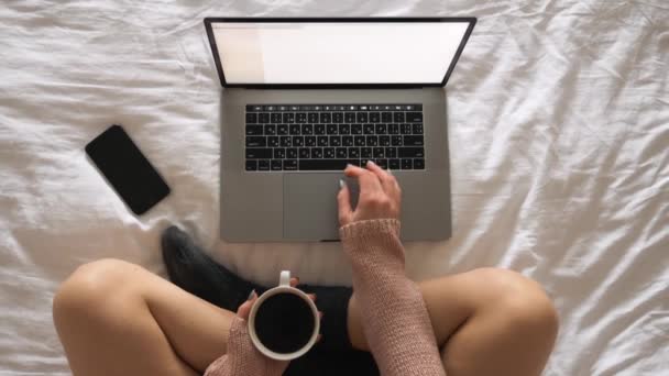 Hermosa joven sentada en la cama por la mañana con una taza de café y latop — Vídeos de Stock